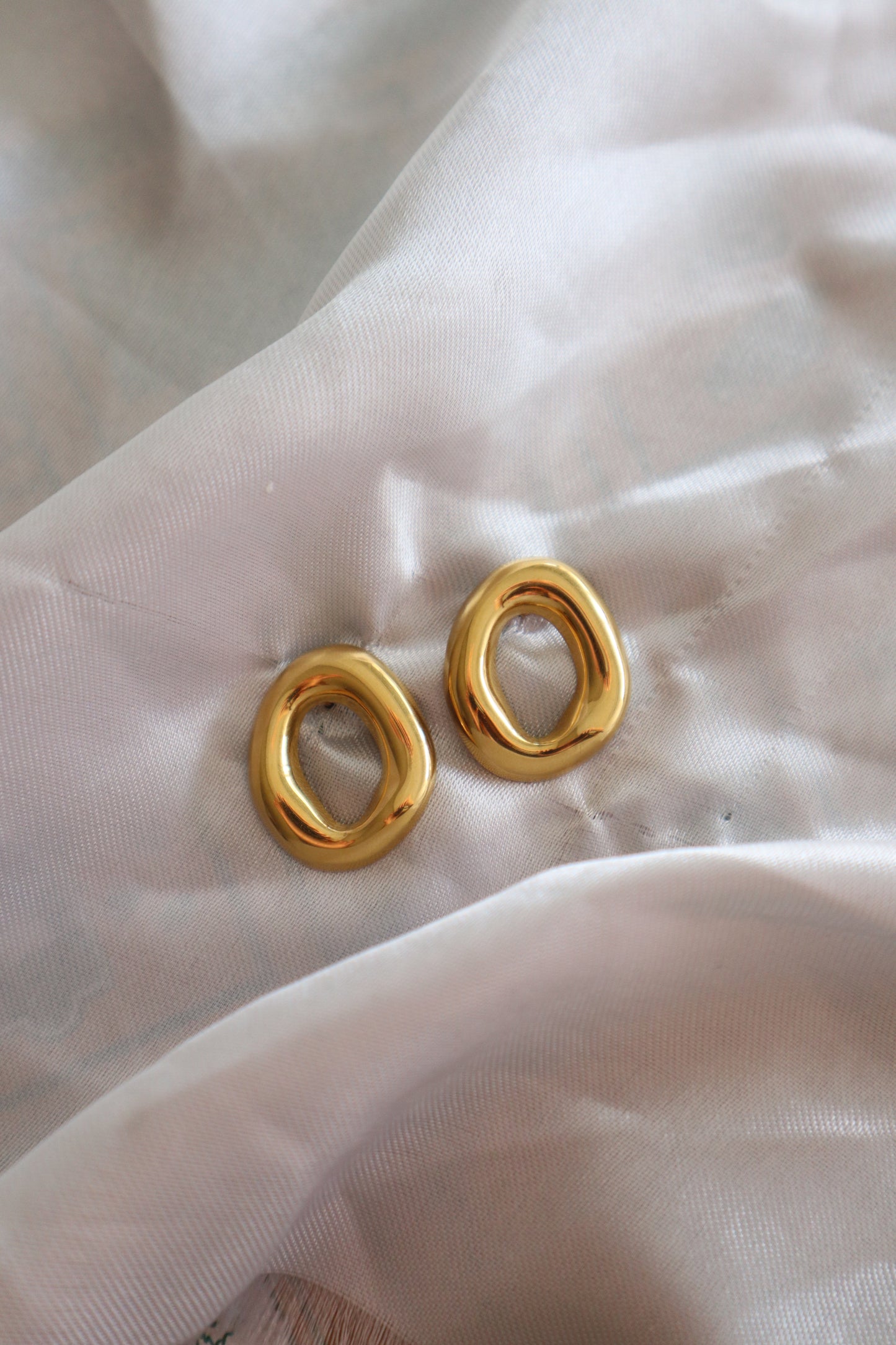Brincos dourados em forma de círculo oco, feitos em aço inoxidável, com design elegante.